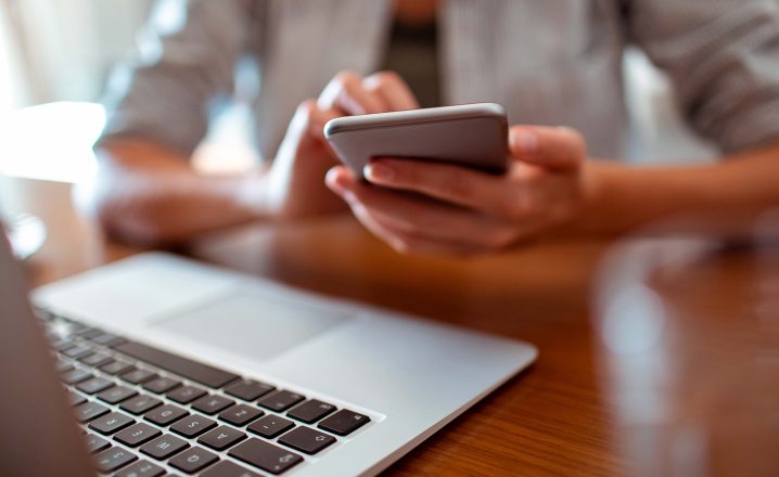 Pictorial representation of a laptop placed on a wooden table with a person's hand holding a mobile phone in the background.